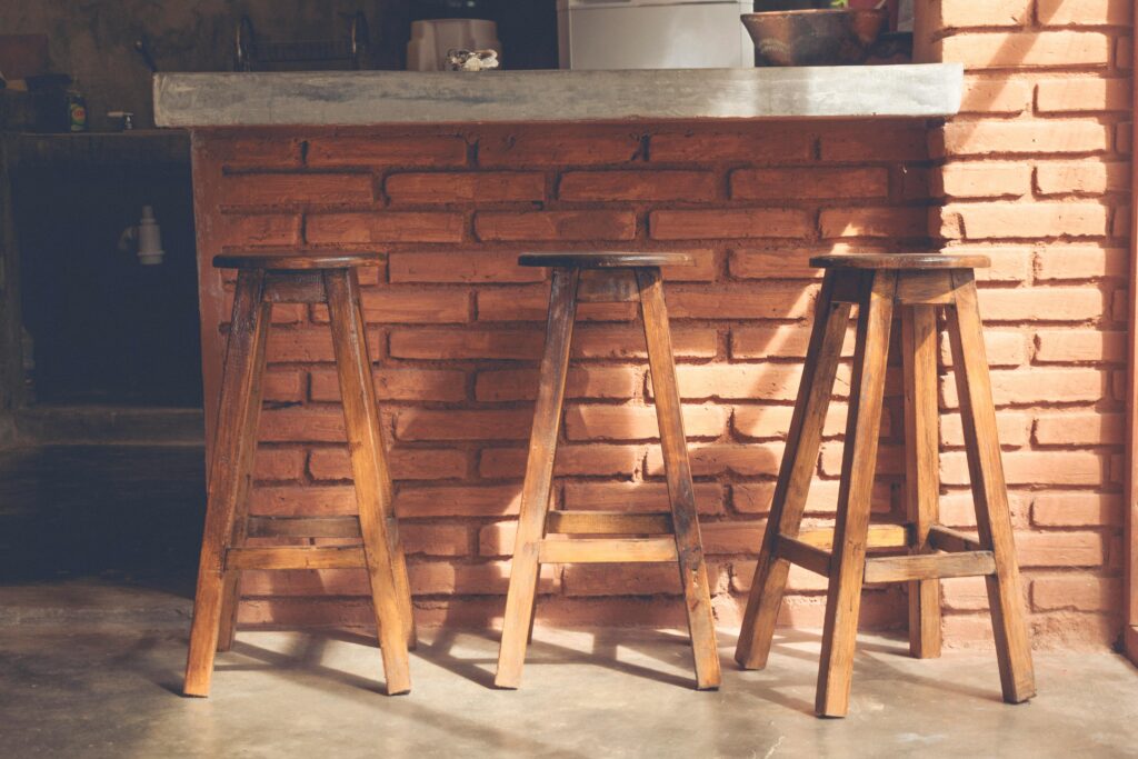 Three wooden bar stools in a rustic kitchen setting with brick walls and natural lighting.